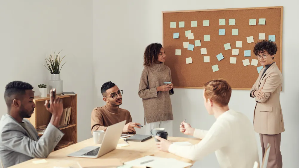 A diverse group of people engaged in a brainstorming session in a modern office. They are gathered around a table with a laptop, while one person stands by a corkboard filled with colorful sticky notes. The scene conveys teamwork and collaborative idea generation