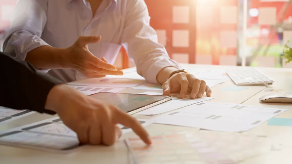 Two people collaborating at a desk, pointing at documents and a tablet, with various papers and design layouts spread out. The scene suggests brainstorming and planning, likely related to the development of a business strategy or plan