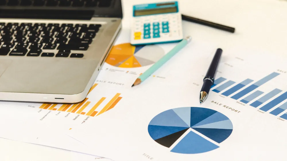 Workspace with a laptop, calculator, pen, and business charts on a desk, including bar and pie charts, representing data analysis and planning. The scene illustrates the process of creating a business plan for a startup