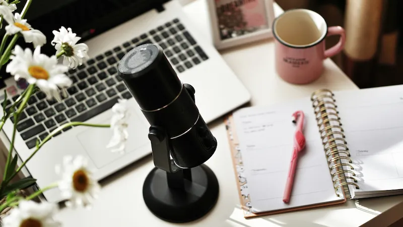 A podcasting microphone sits on a desk alongside a cup of tea and a small vase of flowers, blending a homely yet professional atmosphere. The setup suggests a cozy, comfortable environment for recording while maintaining a level of professionalism, ideal for creative work or content creation