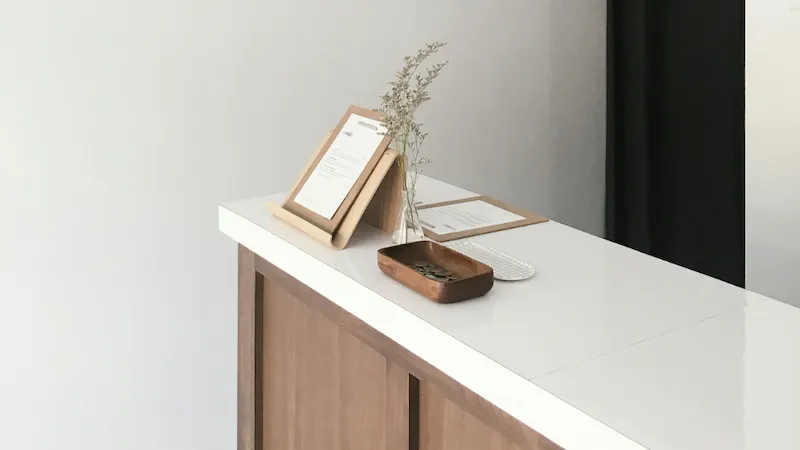 A minimalist reception desk featuring clean lines and a simple setup. The desk includes a clipboard with documents, a vase with dried flowers, and a wooden tray with coins. The modern design highlights a professional, organized, and welcoming workspace