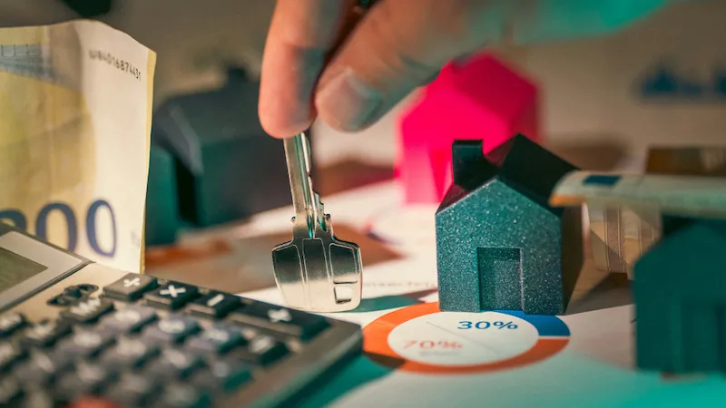 A close-up image showing a hand holding keys near miniature house models on a desk, surrounded by a calculator, euro notes, and charts. The scene symbolizes financial decision-making, investments, and the costs related to upgrading property or workspace