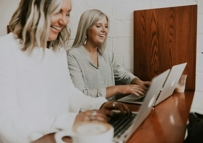 Two women smiling whilst looking at their laptop screens